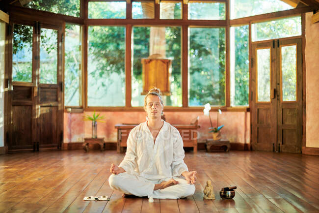 Joven hombre consciente descalzo macho sentado en pose de loto practicando yoga en el suelo cerca del gong bowl y la estatuilla de Buda - foto de stock