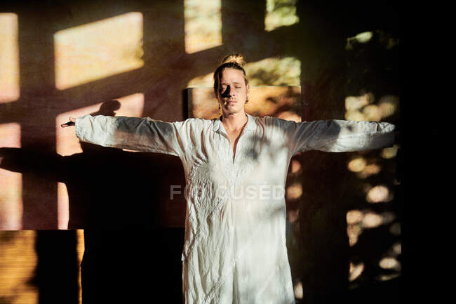 Молодой внимательный мужчина с вытянутыми руками и закрытыми глазами практикующий йогу против стены с оттенками в мягком солнечном свете — стоковое фото