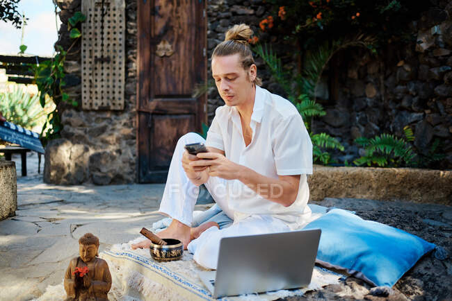 Giovane messaggistica di testo maschile a piedi nudi sul cellulare vicino netbook e statuetta di Buddha nel patio dopo aver praticato yoga — Foto stock