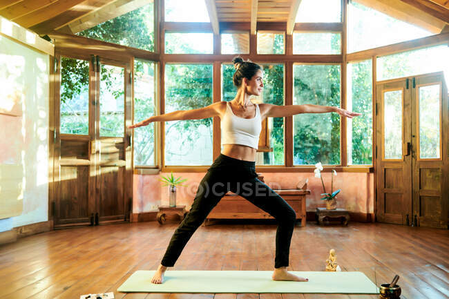Joven hembra descalza en ropa deportiva mostrando la pose Virabhadrasana mientras practica yoga en casa ligera - foto de stock
