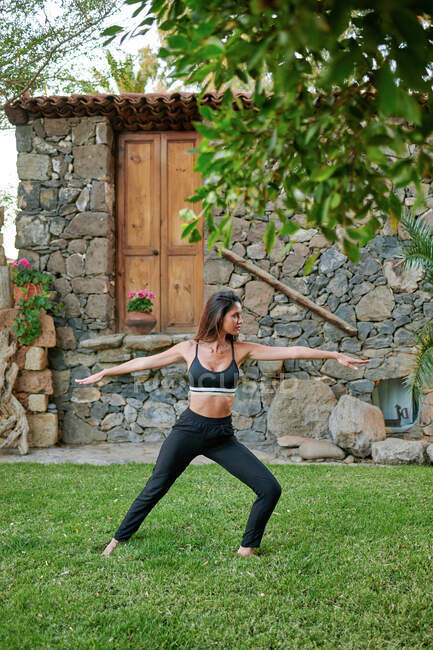 Side view of anonymous flexible female in sports clothes standing in Virabhadrasana pose on lawn in courtyard on summer day — Stock Photo