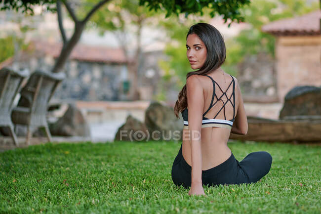 Back view of young fit female in sportswear sitting on lawn while looking at camera over shoulder in courtyard — Stock Photo