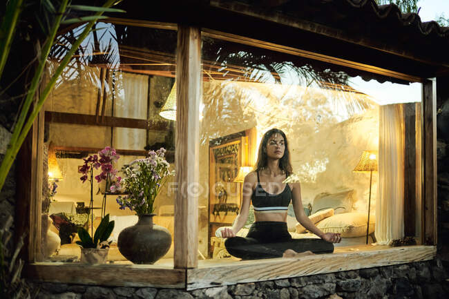 Через окно вид молодой мечтательной женщины с закрытыми глазами медитирующей сидя со скрещенными ногами на стеклянной террасе — стоковое фото