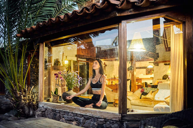 Через окно вид молодой мечтательной женщины с закрытыми глазами медитирующей сидя со скрещенными ногами на стеклянной террасе — стоковое фото