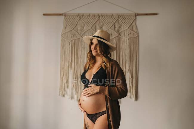 Felice donna incinta in lingerie nera e cardigan commovente pancia e guardando giù in appartamento leggero durante il giorno — Foto stock