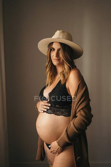 Pensive pregnant woman in black lingerie and cardigan touching belly and looking at camera in light apartment in daytime — Stock Photo