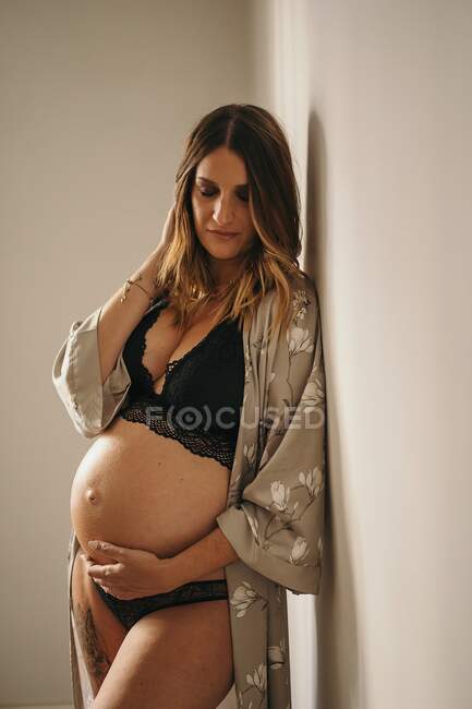 Mujer embarazada pensativa en lencería negra y bata tocando vientre con los ojos cerrados en apartamento de luz en el día - foto de stock