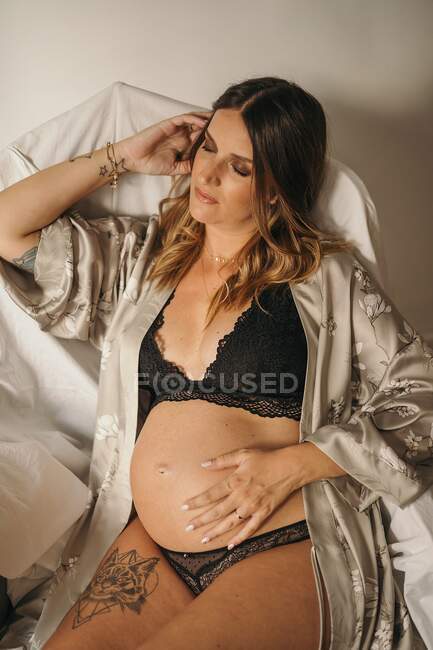 Спокійна вагітна жінка в нижній білизні сидить із закритими очима і торкається живота в світлій кімнаті в квартирі — стокове фото
