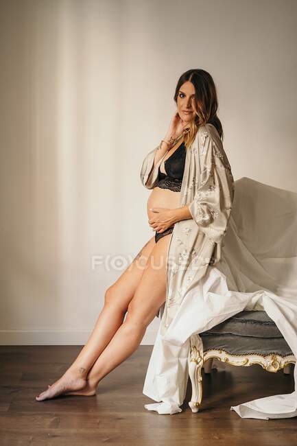 Corpo cheio de mulher grávida encantadora em lingerie elegante e roupão sentado no ângulo do sofá coberto com tecido branco e olhando para a câmera no estúdio de luz — Fotografia de Stock