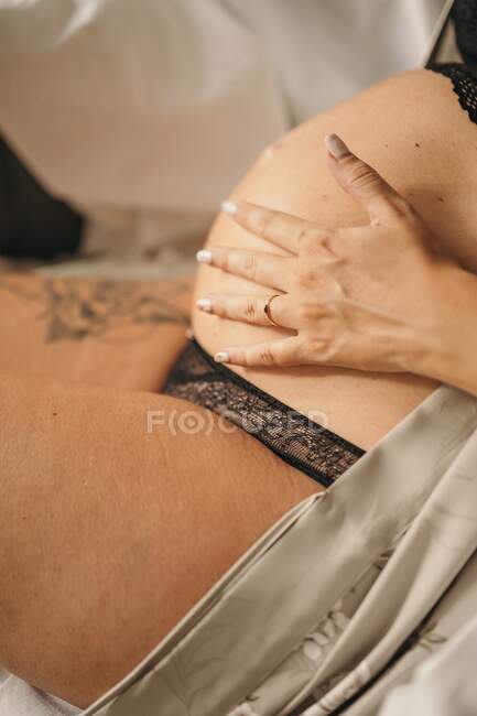 Side view crop of anonymous pregnant woman in stylish lingerie and robe sitting and touching belly — Stock Photo