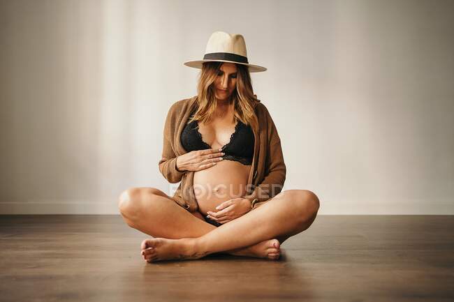 Потрясающая беременная женщина в стильном наряде и шляпе, касаясь живота и глядя вниз, сидя на деревянном полу в квартире — стоковое фото