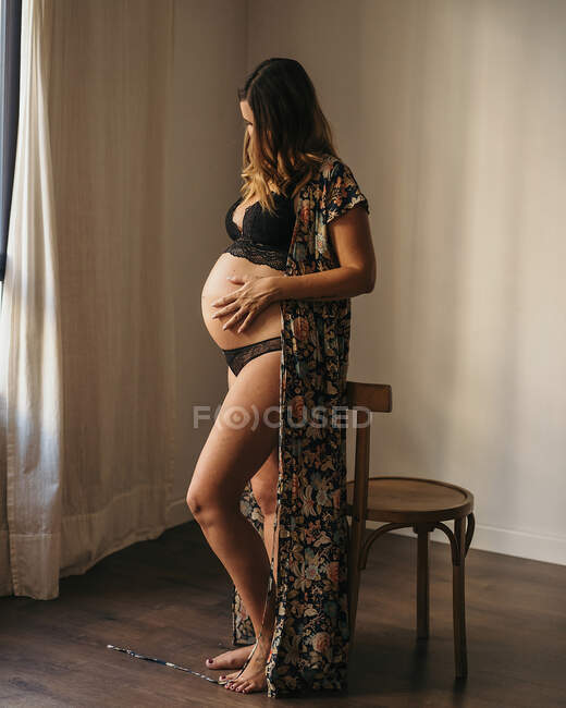 Anonymous pregnant woman in black lingerie and cardigan touching belly and looking down in light apartment in daytime — Stock Photo