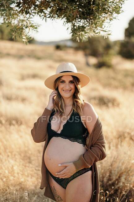 Весела вагітна жінка в нижній білизні капелюха і кардиган стоїть серед сухої трави в полі, розміщеної в сільській місцевості і дивлячись на камеру в сонячний день — стокове фото