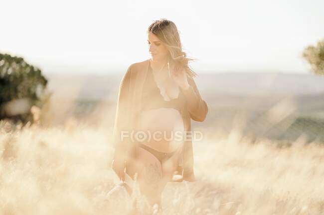 Pensive pregnant woman in lingerie and cardigan standing among dry grass in field placed in countryside and looking down in sunny day — Stock Photo