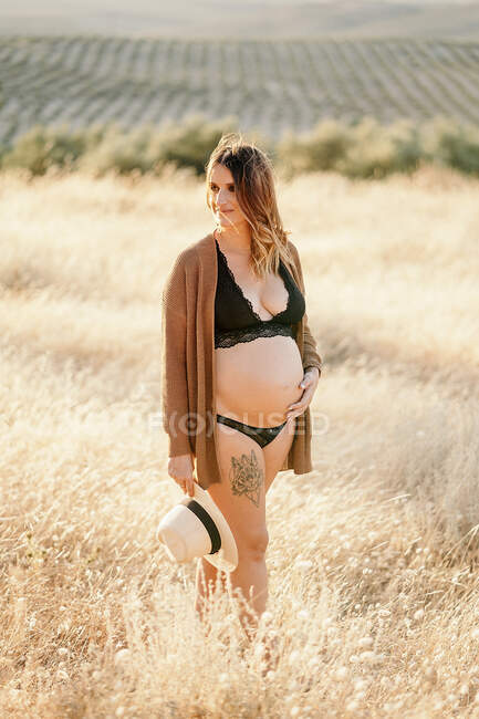 Pensiva mulher grávida em lingerie e casaco de lã em pé entre grama seca no campo colocado no campo e olhando para longe no dia ensolarado — Fotografia de Stock