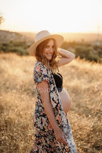 Pensiva donna incinta in lingerie cappello e cardigan in piedi tra erba secca in campo collocato in campagna e guardando altrove nella giornata di sole — Foto stock