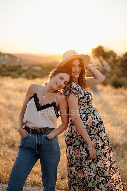 Charming female in trendy outfit leaning head on positive pregnant female while standing in field in countryside and looking at camera in sunny day — Stock Photo