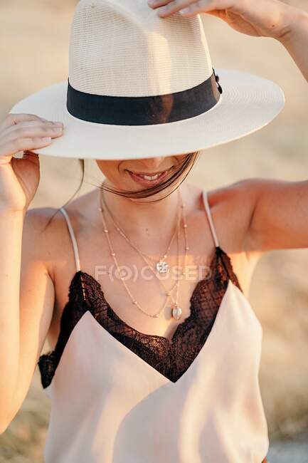 Mujer sonriente con la cara cubierta de pelo oscuro con sombrero y ropa elegante de pie en el campo con hierba seca en el día soleado - foto de stock