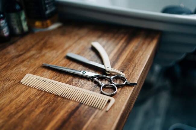 From above scissor and comb near straight razor with sharp metal blades on wooden table in hairdressing salon - foto de stock