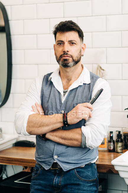 Masculine male stylist holding straight razor with sharp blade while standing in beauty salon in daytime looking at camera — Foto stock