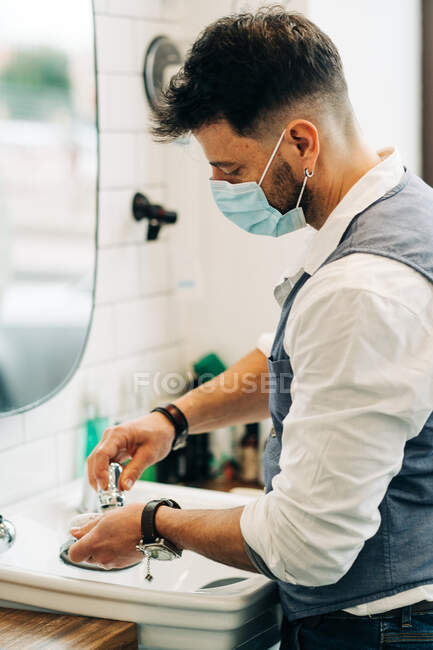 Боковой вид анонимного мастера красоты в стерильной маске, готовящего кисть для бритья с мылом в миске против зеркала в ванной комнате на работе — стоковое фото