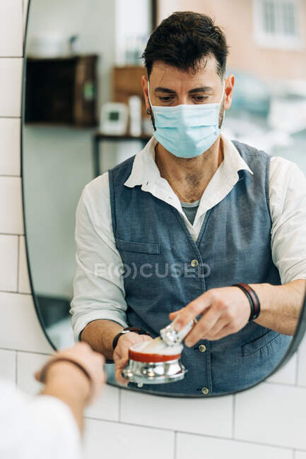 Anonymous male beauty master in sterile mask preparing shave brush with soap in bowl against mirror in bathroom at work - foto de stock