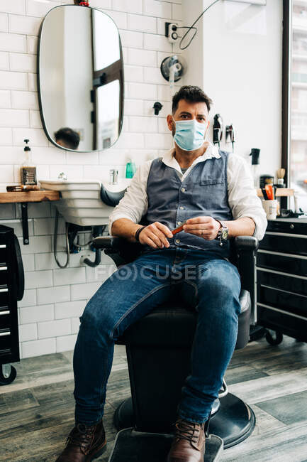 Anonymous male beauty master in stylish wear and face mask sitting in hairdressing salon while looking at camera — Fotografia de Stock