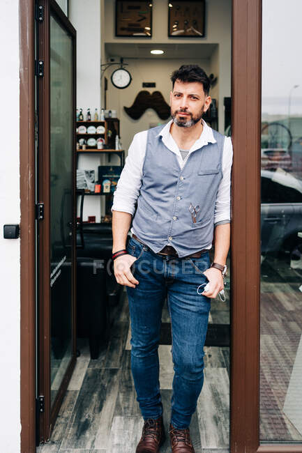 Дорослий бородатий чоловічий майстер краси з руками в кишенях дивиться на камеру біля дверей перукарні — стокове фото