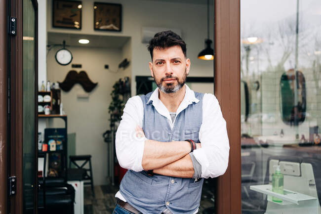 Взрослый бородатый мужчина мастер красоты с скрещенными руками, смотрящий в камеру, стоящую у двери парикмахерской — стоковое фото