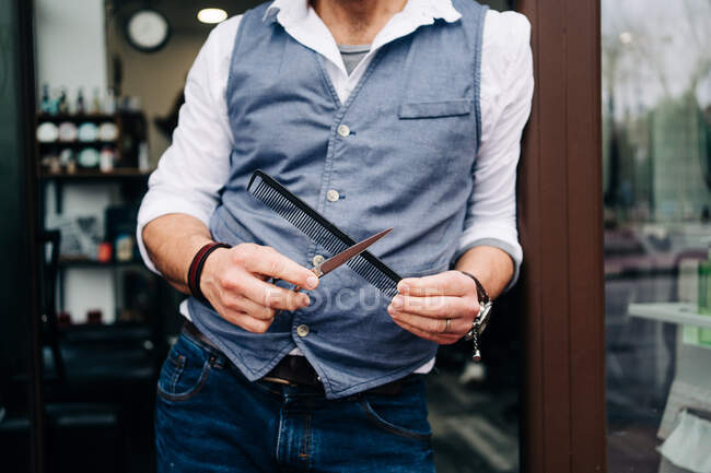 Crop unrecognizable male hairdresser in smart casual apparel with professional grooming tools at entrance door of beauty salon in town - foto de stock