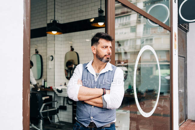 Handsome adult bearded male beauty master with arms crossed looking away standing at door of barbershop — Fotografia de Stock