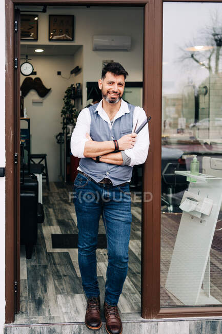 Щасливий усміхнений чоловічий перукар у розумному повсякденному одязі з професійними інструментами для догляду біля вхідних дверей салону краси в місті — стокове фото