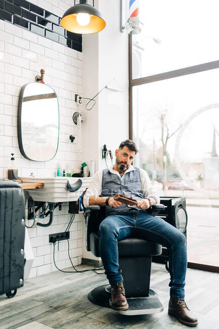 Adult unshaven beauty master in smart casual wear looking at camera under shiny lamp in barbershop — Foto stock