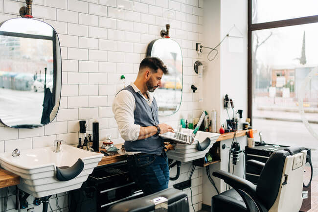Seitenansicht männlicher Friseur surft im Internet auf Netbook im modernen Schönheitssalon gegen Fenster — Stockfoto