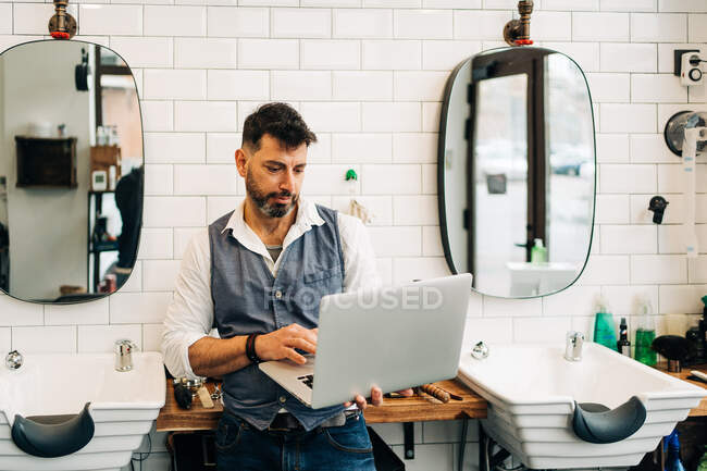 Маскулінний чоловічий перукар переглядає Інтернет на нетбуці в сучасному салоні краси проти вікна — стокове фото