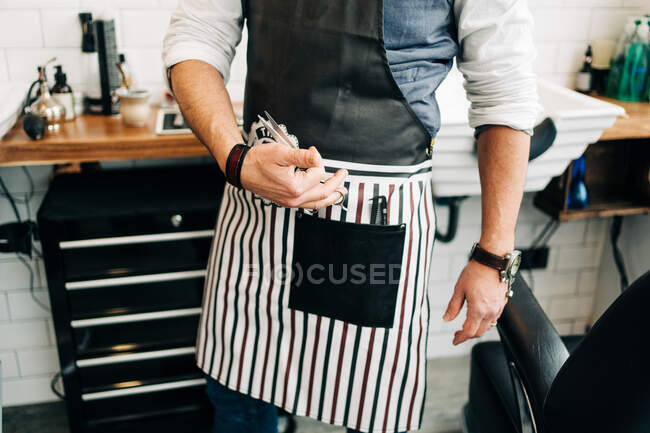 Crop unrecognizable male barber with scissors and comb in striped apron in hairdressing salon - foto de stock