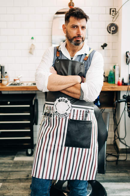 Self assured adult bearded male hairstylist in apron looking at camera with folded arms in barbershop - foto de stock