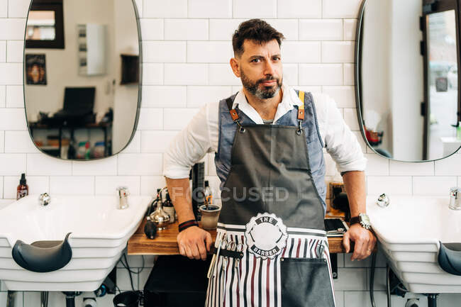 Selbstbewusster erwachsener bärtiger männlicher Friseur in Schürze blickt im Friseurladen in die Kamera — Stockfoto