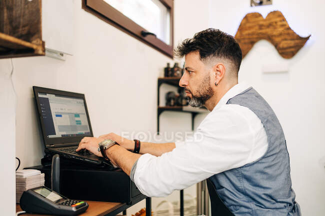 Vue latérale du coiffeur masculin concentré travaillant sur netbook avec programme ouvert à l'écran dans un salon de beauté près de la caisse enregistreuse jusqu'au lecteur de carte — Photo de stock