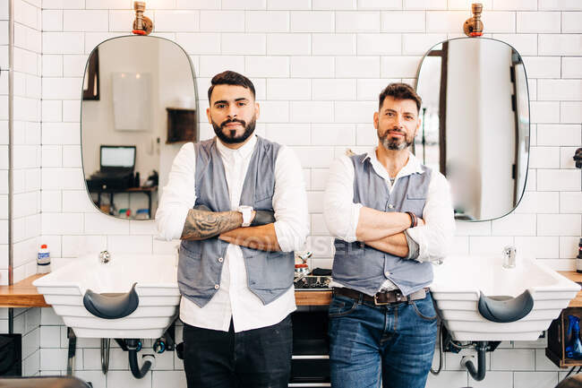Adult self confident hairdressers in smart casual apparel looking at camera with folded arms in barbershop — Stock Photo