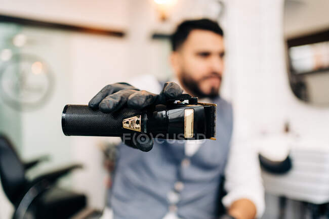 Masculine male hairdresser in glove demonstrating professional electric clipper in barbershop on blurred background — Fotografia de Stock