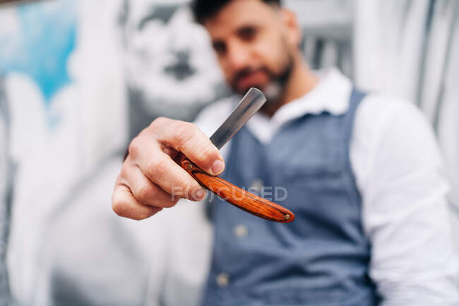Crop masculine male stylist demonstrating straight razor with sharp blade in beauty salon in daytime - foto de stock