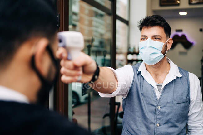 Male hairdresser in sterile mask measuring temperature of crop anonymous colleague with infrared thermometer at door of barbershop — Stock Photo