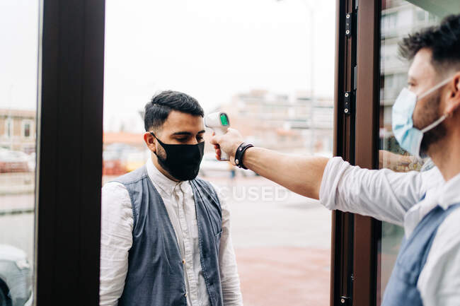 У стерильній масці, що вимірює температуру колеги з інфрачервоним термометром на дверях перукарні. — стокове фото