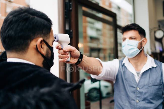 Male hairdresser in sterile mask measuring temperature of crop anonymous colleague with infrared thermometer at door of barbershop — Fotografia de Stock