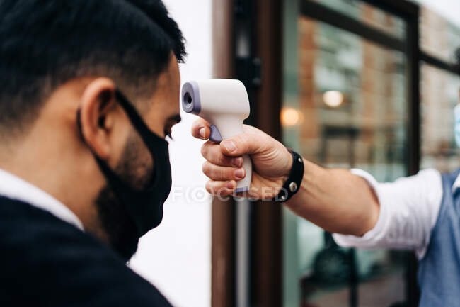 Culture anonyme salon de coiffure masculin en masque stérile mesure de la température de collègue avec thermomètre infrarouge à la porte du salon de coiffure — Photo de stock