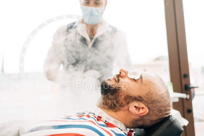 Crop anonymous beauty master in mask near adult bearded male client with cotton pads on closed eyes during vapor procedure in barbershop — Foto stock