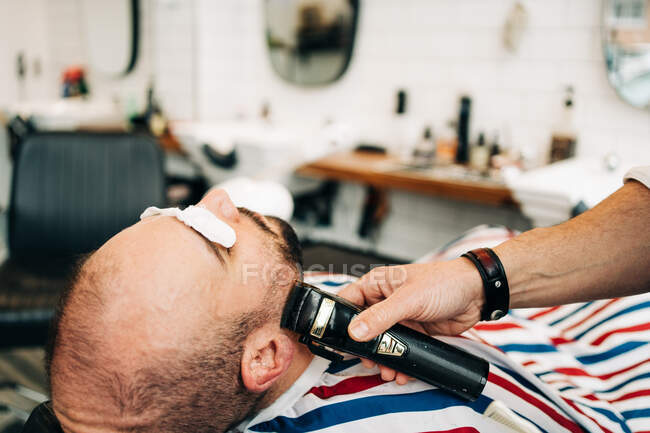 Crop anonymous master trimming beard of masculine man with electric machine in hairdressing salon - foto de stock