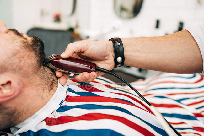 Crop anonymous master trimming beard of masculine man with electric machine in hairdressing salon — Fotografia de Stock