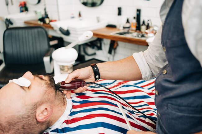 Crop anonymous master trimming beard of masculine man with electric machine in hairdressing salon - foto de stock